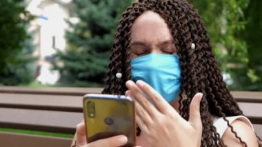 Koruyucu maske takan neşeli, genç bir kadın parkın dışında akıllı telefon kullanıyor.