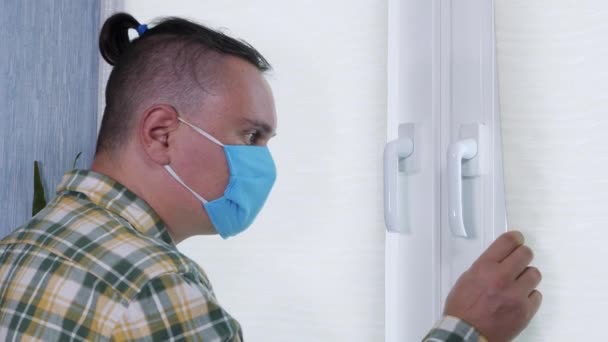 Hombre triste adulto con máscara protectora mira por la ventana durante la cuarentena — Vídeos de Stock