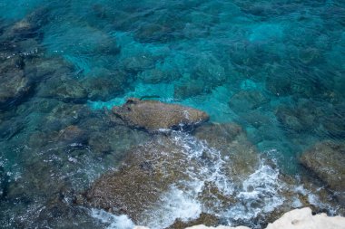 Kıbrıs Rum Kesimi 'nin Ayia Napa kentindeki kayalık Akdeniz kıyı şeridinin çarpıcı manzarası. Dalgalar, adanın doğal güzelliğini ve egzotik cazibesini gözler önüne seren parlak bir gökyüzü altında taştan kıyılara çarpar..