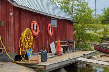 A rustic wooden boathouse with weathered walls, featuring a hanging hose, lifebelts and various tools neatly arranged, Baltic sea, Finland