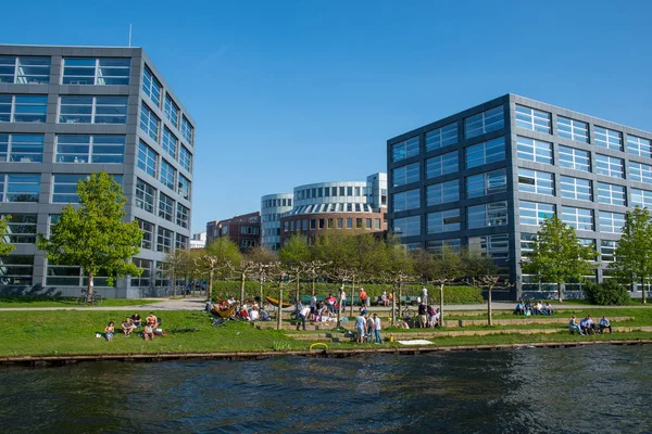 Personas disfrutando de un día soleado a orillas del río Spree con edificios de oficinas en el fondo — Foto de Stock