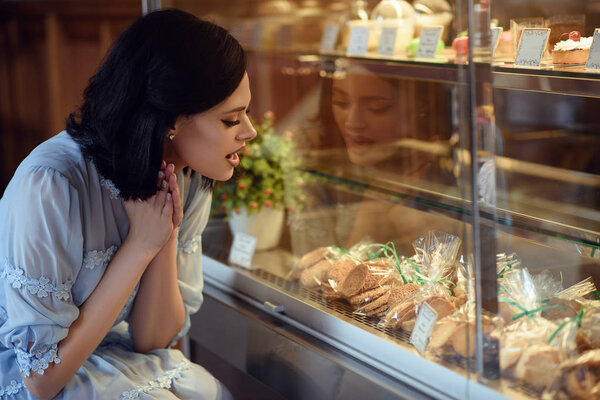 Portrait of young beautiful girl looking at the showcase with cakes and biscuits with excitement on her face.