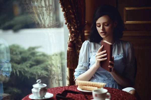 Молодая красивая леди сидит за столом в уютном кафе с книгой в руках. Чашка кофе и вафельные рулетики на столе Лицензионные Стоковые Фото