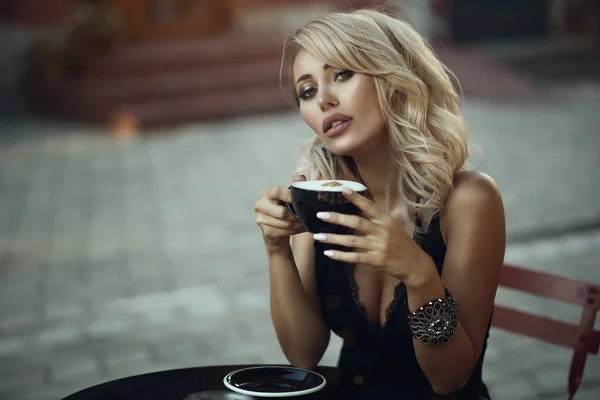 Portrait of gorgeous elegant blond woman sitting at the table in the nice street cafe holding a cup with frothy latte. — Stock Photo, Image