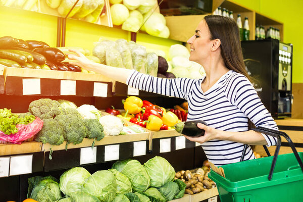 Smiling caucasian brunette woman with basket trying to choose vegetables in grocery