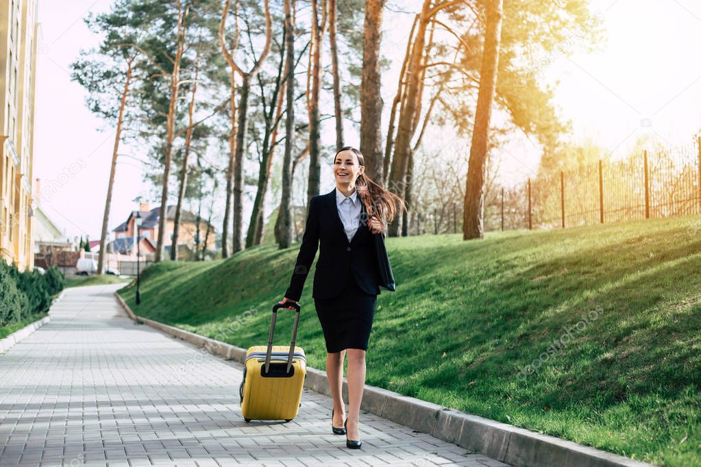 Young caucasian brunette businesswoman in suit with yellow suitcase outdoors. Business trip concept.