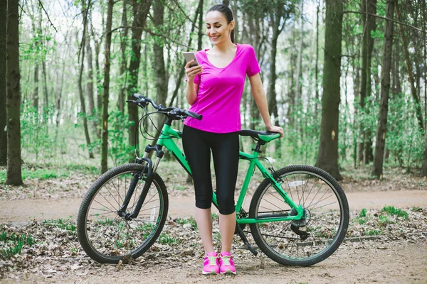 Sporty woman with phone and mountain bicycle in forest