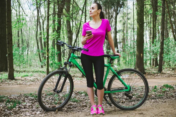 Sporty woman with phone and mountain bicycle in forest