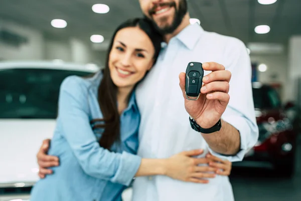 Happy couple hugging and showing key standing on car background in dealership, crop