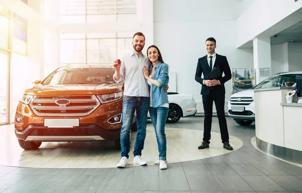 Happy young family standing with car key in dealership, male manager and car on background, full length