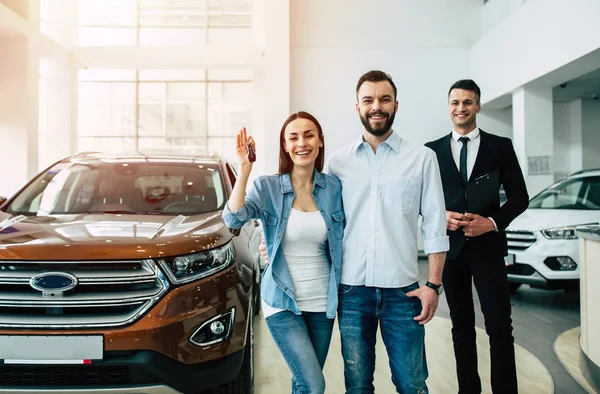 Happy young family standing with car key in dealership, male manager and car on background
