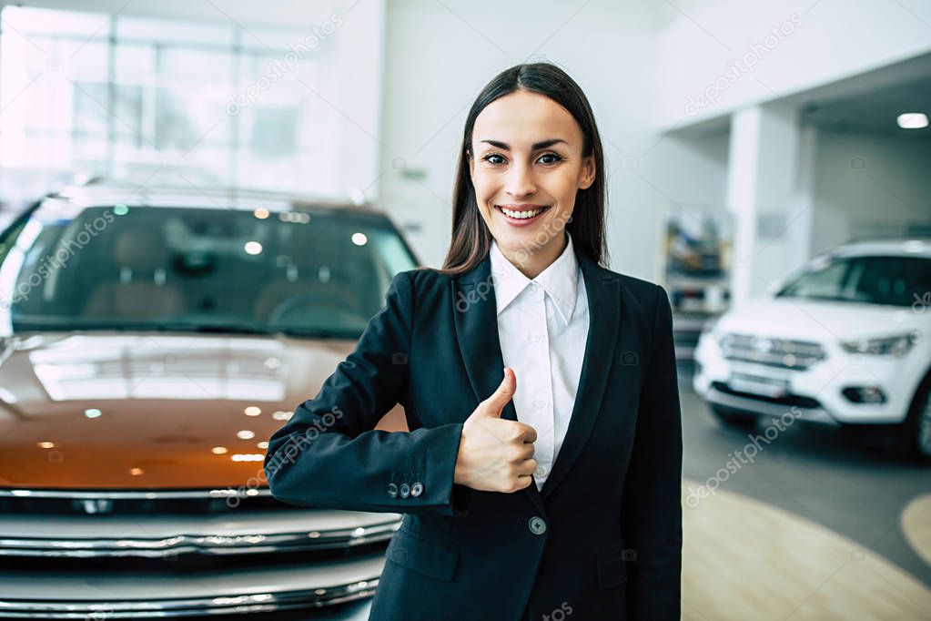 portrait of cheerful smiling businesswoman in black suit showing thumb up standing on dealership cars background