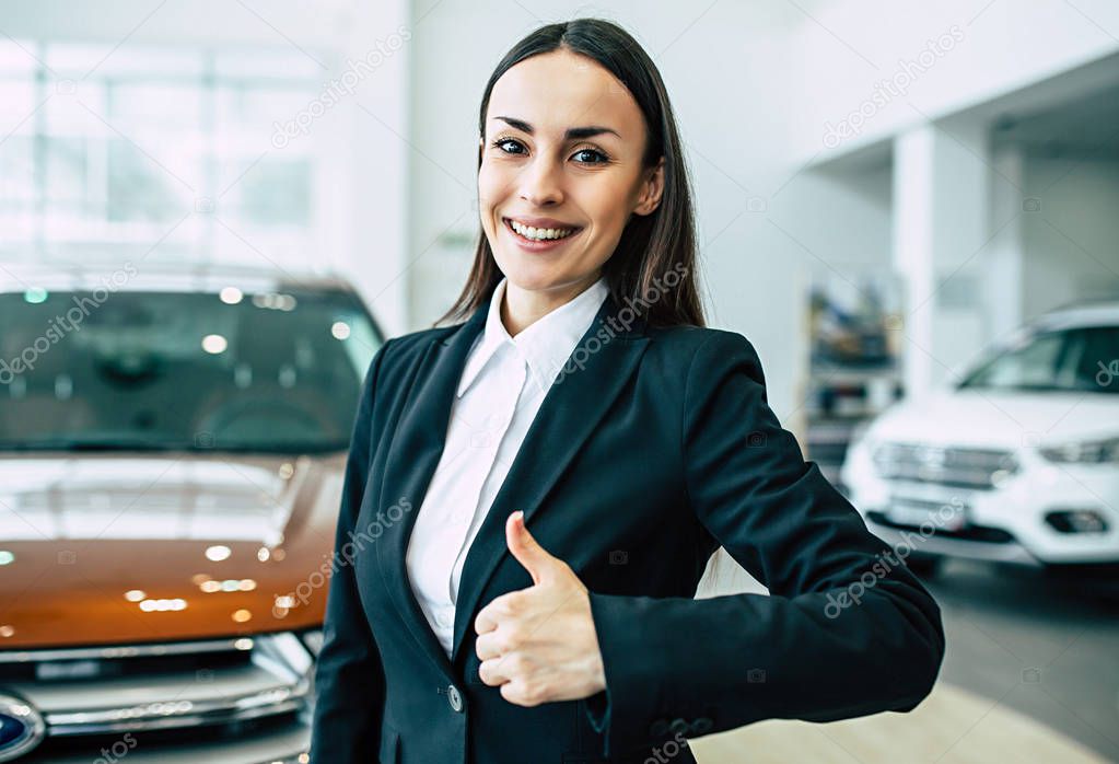 portrait of cheerful smiling businesswoman in black suit showing thumb up standing on dealership cars background