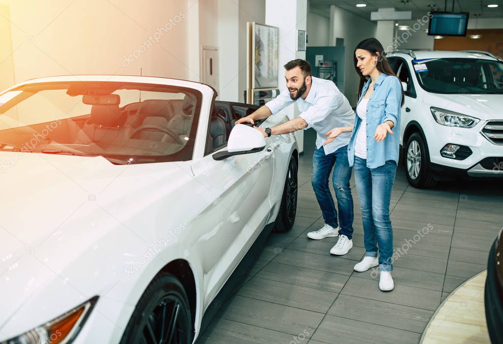 Happy young couple choosing new car in dealership, exciting man touching car
