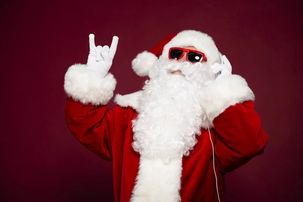 portrait of man in Santa Clause costume listening to music at headphone and showing rock sign gesture on red background, Christmas and New year concept