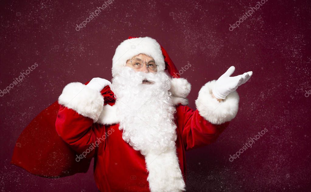 portrait of man in Santa Clause costume with gift sack on back pointing with hand aside on red background, Christmas and New year concept  