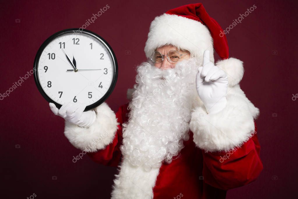 portrait of man in Santa Clause costume holding white clock and showing attention sign on red background, Christmas and New year concept 