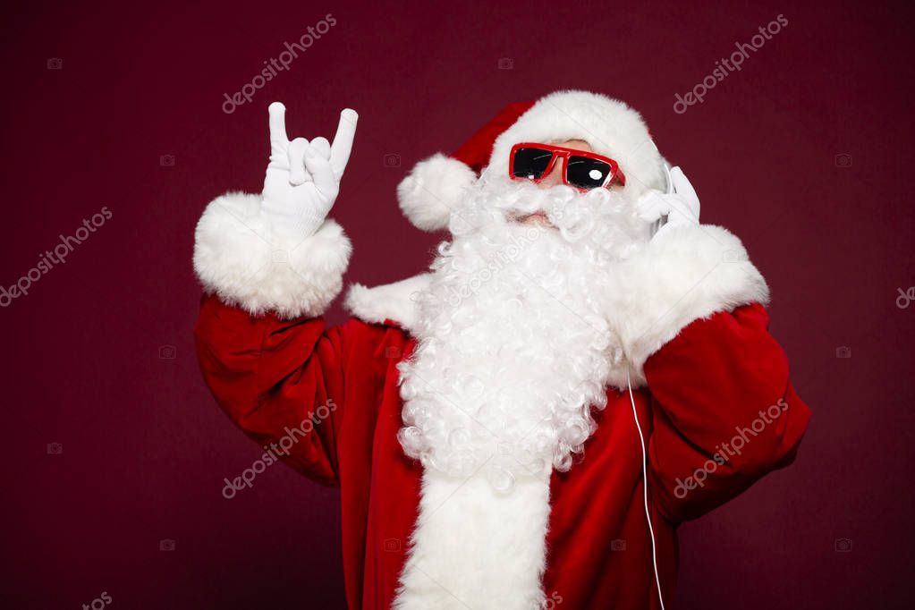 portrait of man in Santa Clause costume listening to music at headphone and showing rock sign gesture on red background, Christmas and New year concept  