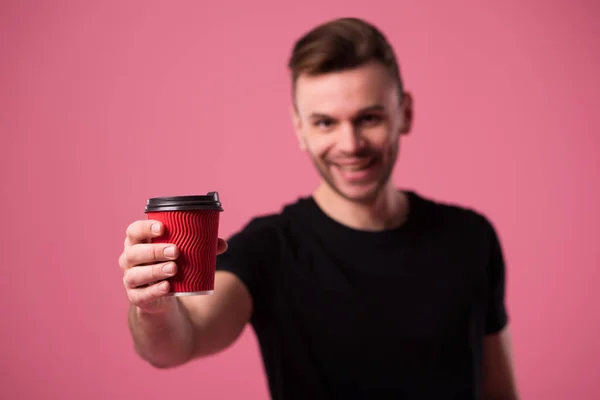 Portrait of handsome smiling man in black t-shirt with cup of coffee in hand on pink background