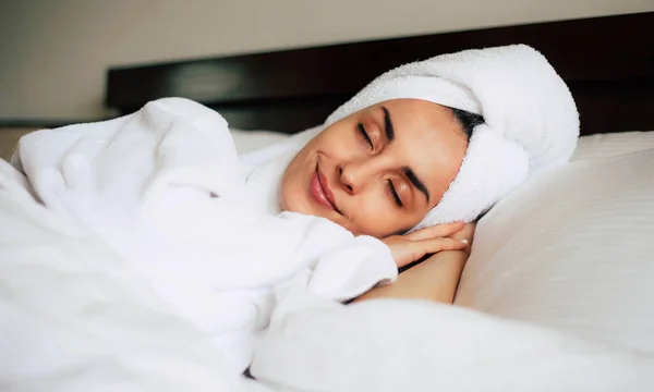 Middle day nap. Cute young girl is having a relaxing nap after shower in cozy double bed with bright white bedclothes. Her skin is tanned, eyebrows are nicely shaped and her smile is pleasant.