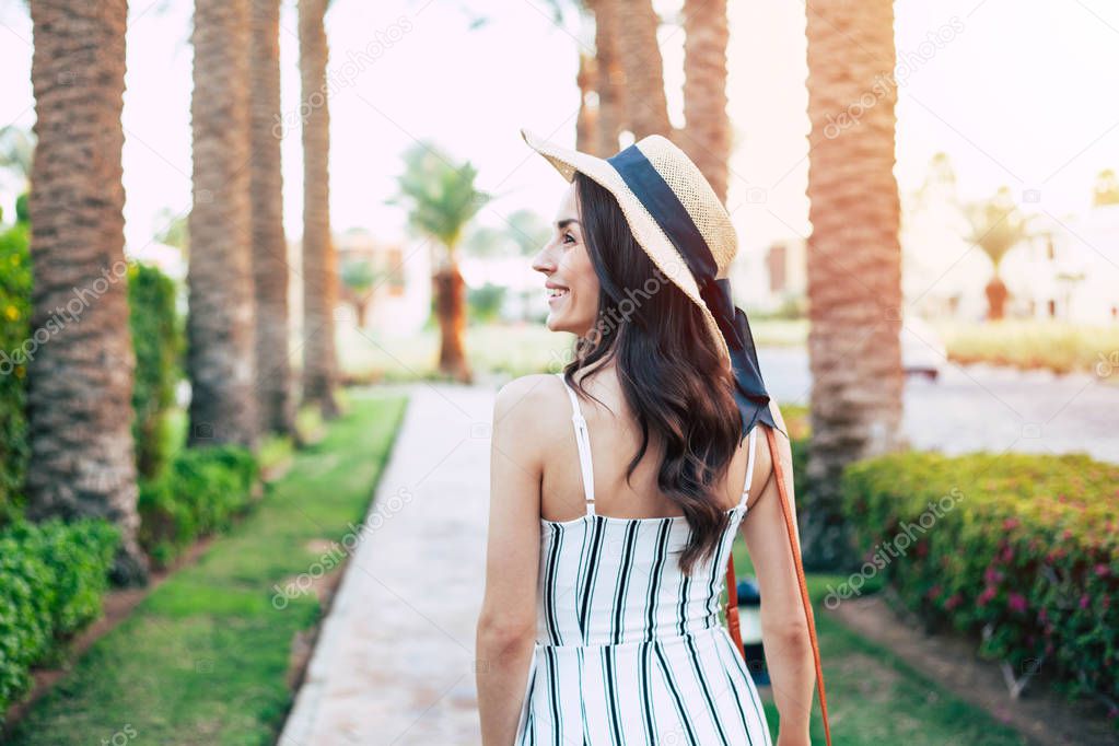Affection to the dreams. Delightful girl with smooth sun lights on her brown hair between palms and bright green bushes wearing black & white jumpsuit, red bag and beige hat with black bow on it.