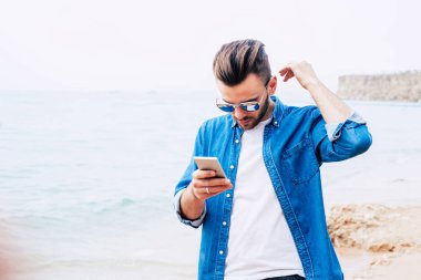 Yakışıklı adam altın kumları ve berrak mavi su yakınında deniz kıyısında saçları düzeltmeye ve onun nişanlısı için onun smartphone üzerinde konuşuyor.