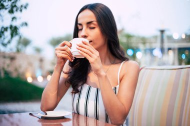 Holibobs. Memnun kız uzun hazel-kahverengi saçlı. Parlayan tabaklanmış deri ve zarif eller tatma lezzetli çayını yeşil ağaçlar ve akşam ışıklar arasında