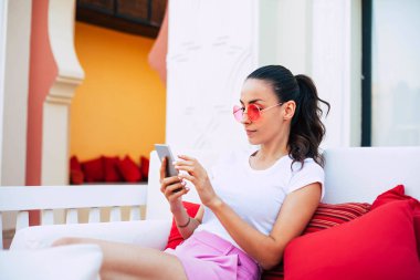 Küçük bir cennet. Altın rengi kırmızı yastık lounge'dan oturan ile at kuyruğu beyaz t-shirt ve pembe şort ile kırmızı güneş gözlüğü eşliğinde rahat kadınla çerçeveli.