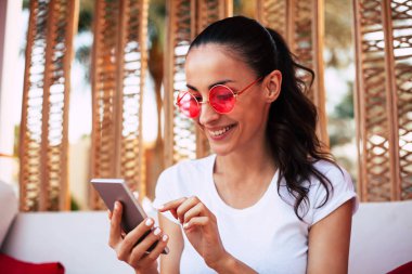 İleti çıkılıyor. Sevimli genç kız beyaz t-shirt ve ahşap dekorasyon ve beyaz kanepe önünde yuvarlak kırmızı güneş gözlüğü telefonunu otel lobisinde kullanıyor.