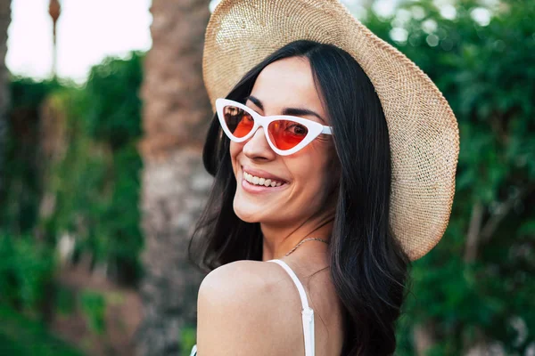 Travel bug. Gorgeous girl with long dark hair, brilliant smile and hazel eyes wearing camel hat and cat shaped sunglasses in front of deep green bushes and a palm tree.