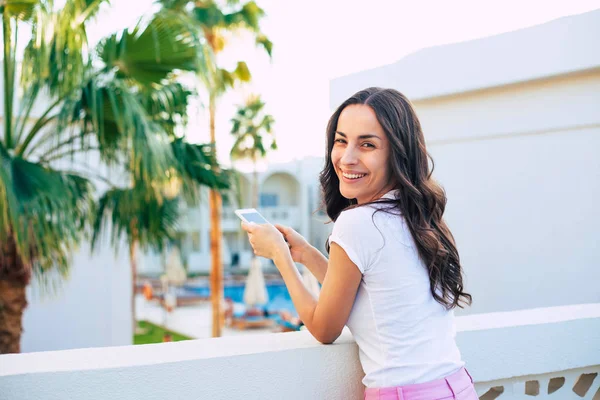 The serenity of the sky. Superb girl with a long curly hair and stunning sincere smile near the palm tree,white building and blue swimming pool wearing light t-shirt and pale pink shorts.