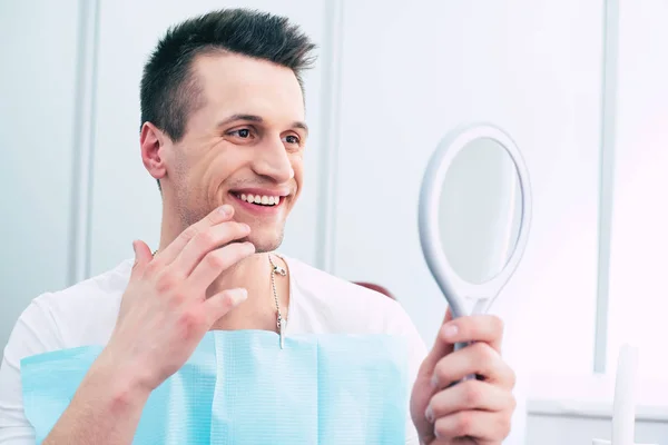 Nice and neat. A photo of pleased man in a dentist cabinet with a mirror in his hand which hi is using for observing the final result of the treatment and he looks absolutely satisfied.