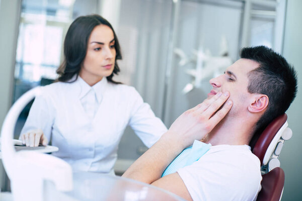 In need of a medical help. A man in a dental chair near the professional dentist is showing that he has a horrible tooth pain with his gestures and asking for a treatment.