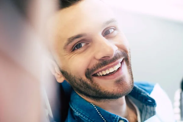 Owner of a smile. Hilariously handsome man clothed in a denim shirt is sitting in a dental hair showing to the doctor his new eye catching healthy smile.