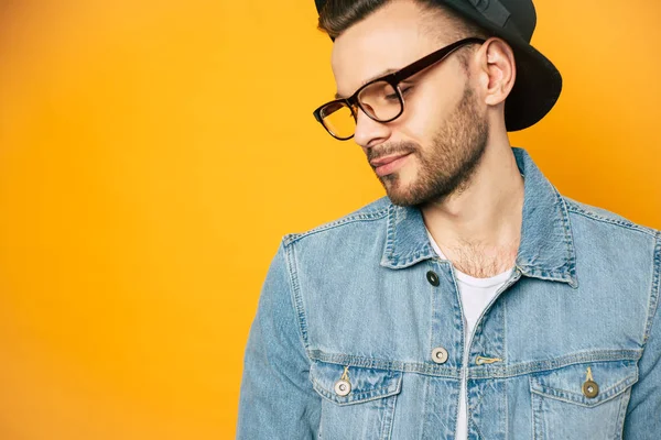 Zen-like calm. A man in a calm mood with a slight smile is wearing a denim jacket, white t-shirt, simple glasses and black hat over his shiny dark brown hair.