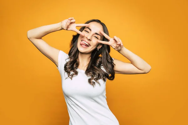 Find peace. Gorgeous young girl next to the orange-colored background with splendid smile all over her face and peace signs which she shows with her hands.