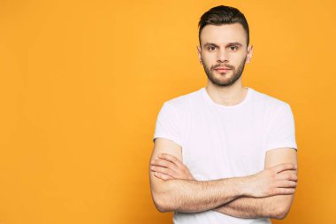 Ciddi bir yüzü olan sakin bir adam beyaz tişörtlü baharatlı-turuncu arka planın önünde duruyor ve elleri göğsüne geçti.