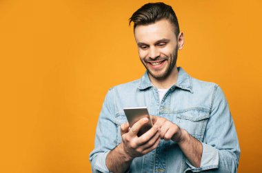 Kot giyen genç modern stil sahibi adam sarı arka planda dururken elinde akıllı telefon kullanıyor.
