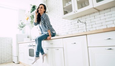 Güzel sevimli gülümseyen kadın evde mutfakta akıllı telefon kullanıyor.