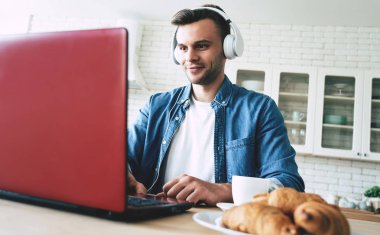 Mutlu sabahlar. Güzel gülümseyen adam evde mutfakta otururken dizüstü bilgisayar kullanıyor ve içecekler kahve veya çay