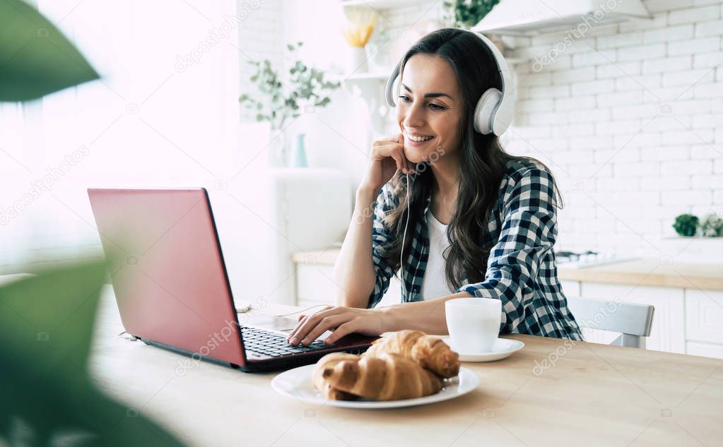 Cute young woman in headphones is watching film on laptop on the kitchen at home