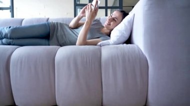 Dinlenmek için zamanın var. Mutlu, sevimli genç bir kadın evdeki kanepede yatarken elinde akıllı telefon kullanıyor..