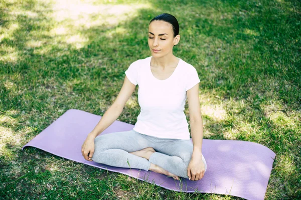 Female Yoga at park. Young brunette woman in lotus pose sitting on green grass in park. Concept of calm and meditation.