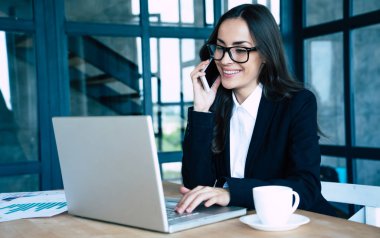 Modern ofiste otururken aynı anda akıllı telefon ve dizüstü bilgisayar kullanarak tam takım giyinmiş büyüleyici iş kadınları