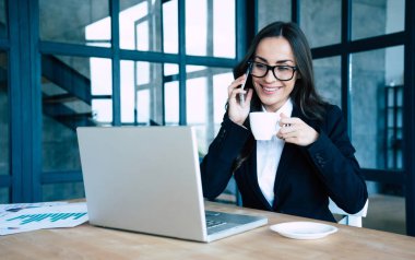 Modern ofiste otururken akıllı telefon ve dizüstü bilgisayar kullanarak tam takım elbise giymiş Charming iş kadınları