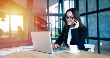 Modern ofiste otururken akıllı telefon ve dizüstü bilgisayar kullanarak tam takım elbise giymiş Charming iş kadınları