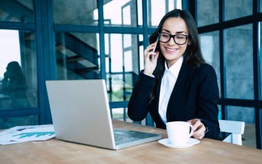Modern ofiste otururken akıllı telefon ve dizüstü bilgisayar kullanarak tam takım elbise giymiş Charming iş kadınları