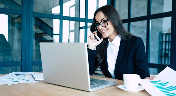 Modern ofiste otururken akıllı telefon ve dizüstü bilgisayar kullanarak tam takım elbise giymiş Charming iş kadınları