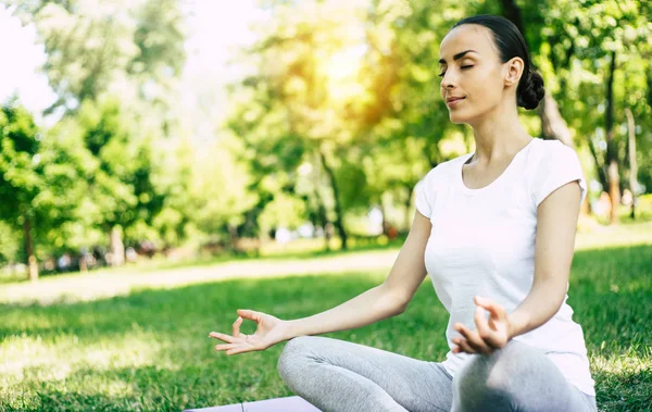 Female Yoga at park. Young brunette woman in lotus pose sitting on green grass in park. Concept of calm and meditation.