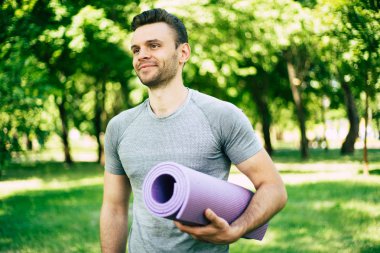 Yakışıklı sportif, ince ve güçlü sporcu veya yoga eğitmeni şehir parkı arka planda elinde yoga mat ile uzak görünüyor